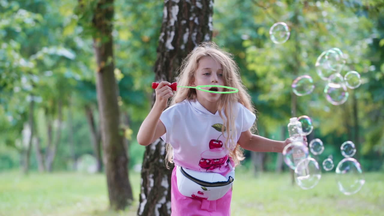 Portrait of a beautiful little girl blowing soap bubbles near the tree. Long-haired child having fun with bubble blower outdoors. Happy childhood.