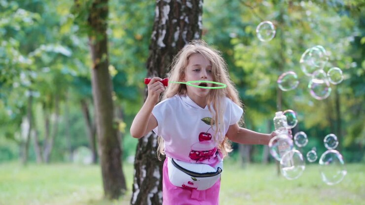 Portrait of a beautiful little girl blowing soap bubbles near the tree. Long-haired child having fun with bubble blower outdoors. Happy childhood.