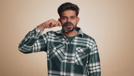 Indian man holding magnifier glass on teeth looking at camera showing funny smiling face mouth
