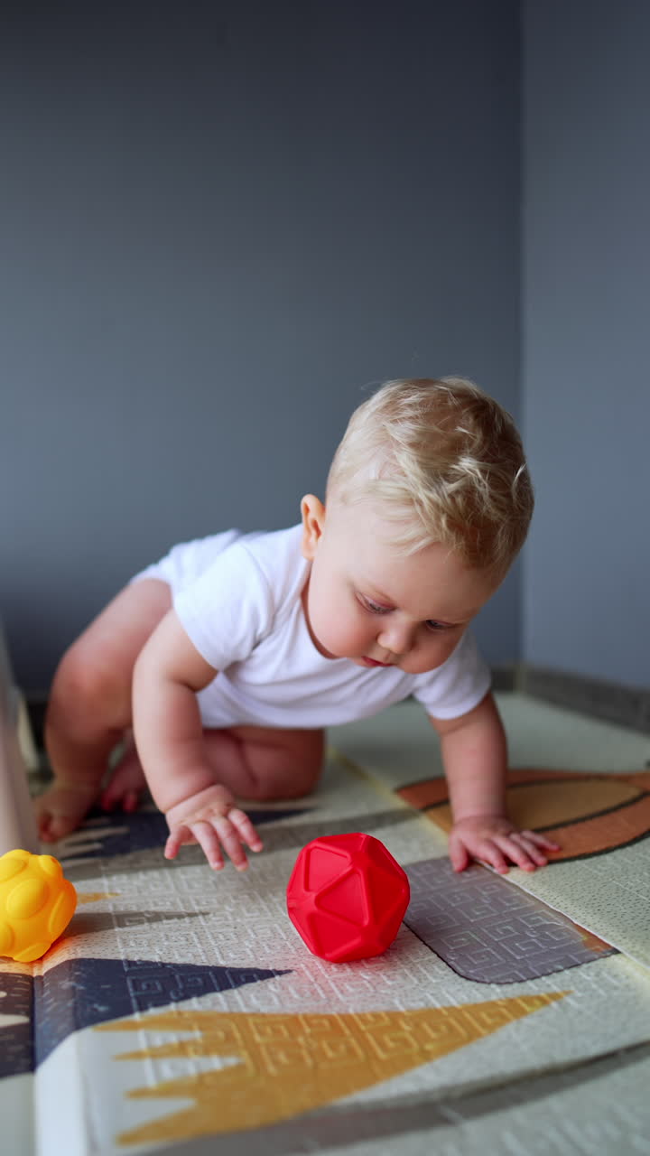 Beautiful blond Caucasian baby holding a toy. Curious kid drops on the floor, crawls and grabs another toy. Vertical video.