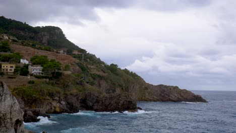 Waves-hitting-the-base-of-the-mountains-with-a-forest-and-some-houses-in-the-background