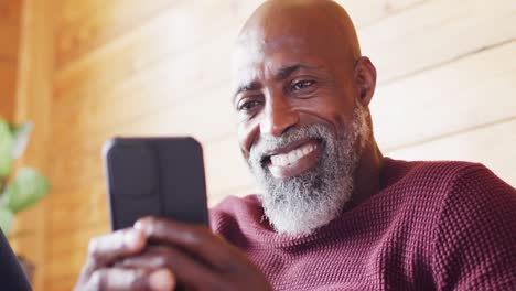 Happy-senior-african-american-man-spending-time-in-log-cabin,-sitting-on-sofa-and-using-smartphone