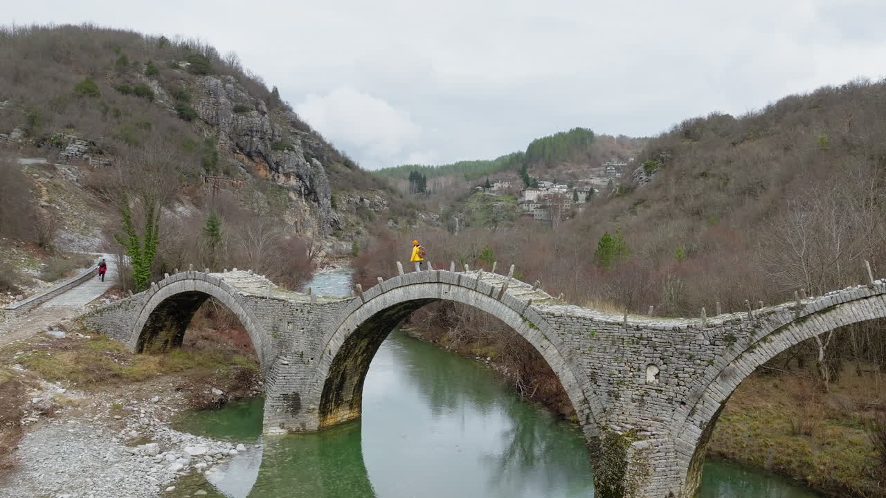 Droneudsigt over Kalogeriko (Plakidas) stenbro i Epirus, Grækenland, med en person der krydser over en smaragdgrøn flod.