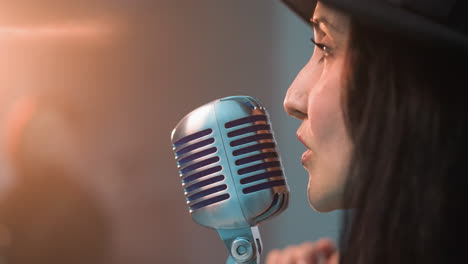 close side view of a woman in a black hat blowing a kiss to a vintage microphone with a smile, against a blurred background with warm lighting, capturing an intimate and emotive moment