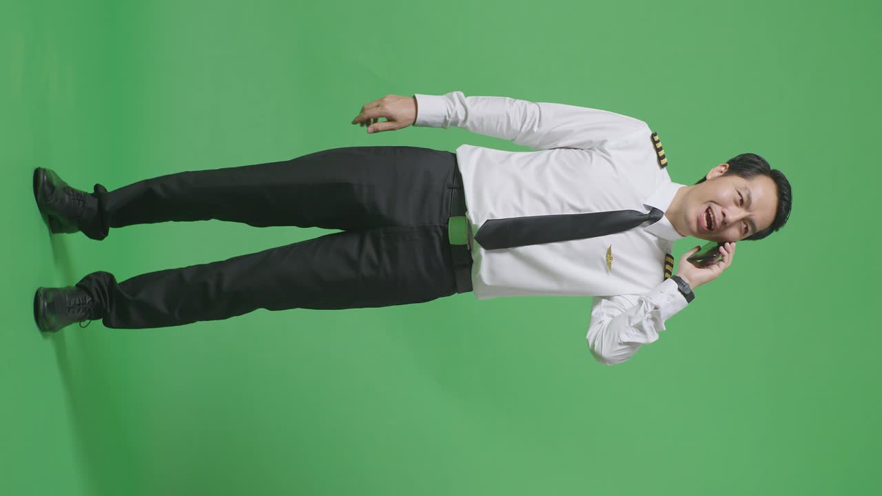 녹색 스크린 배경 스튜디오에 서 있는 동안 스마트폰으로 이야기하고 주변을 둘러보는 아시아 남성 조종사의 몸이 가득 차 있습니다.