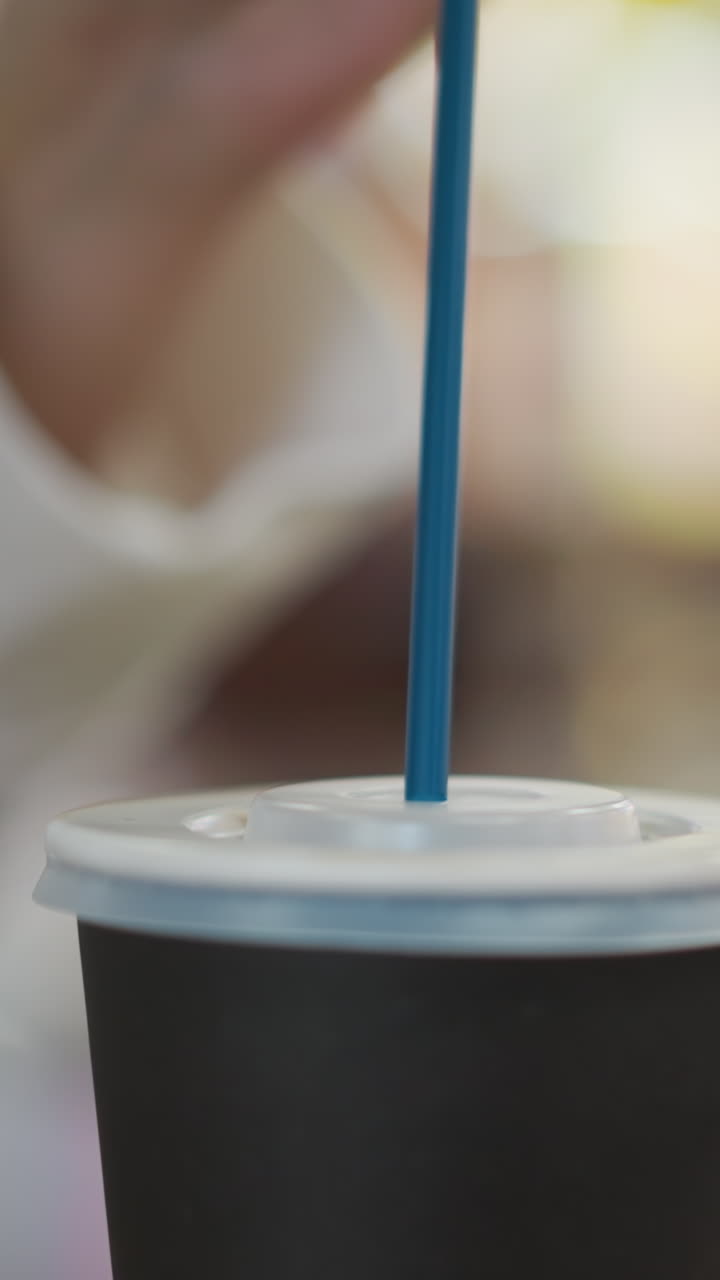 Close-up of hand gently inserting a black straw into a takeaway tea cup lid, creating a minimalist scene with a soft blurred background and bokeh lights
