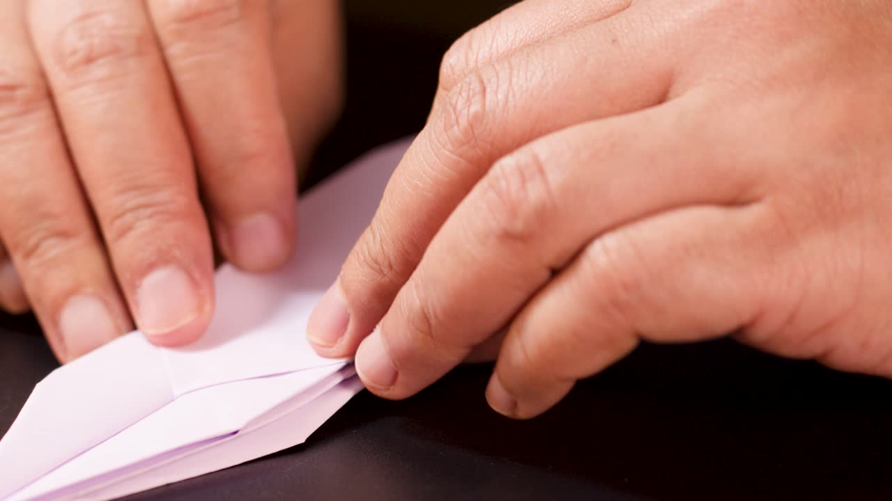 Close-up hands folding purple paper into origami ship, soft lighting, steady camera, dark background