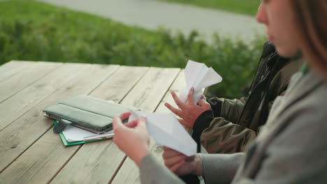 close up rear view of person holding folded paper kite while seated at wooden table outdoors, showing creative crafting activity with casual atmosphere and focus on handmade design