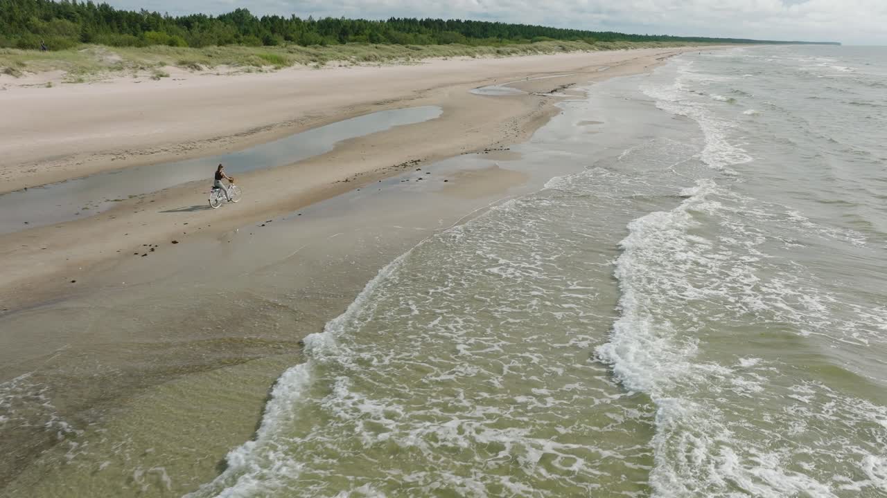 Veduta aerea con una giovane ragazza dai capelli lunghi che va in bicicletta sulla spiaggia sabbiosa, giornata di sole, spiaggia di sabbia bianca, concetto di stile di vita attivo, ampia ripresa aerea con drone in movimento in avanti