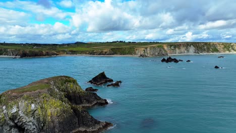 drone flying to land over little island and dark rocks with deep emerald green seas and dramatic puffy clouds coastline establishing shot of the dramatic waterford coastline in ireland