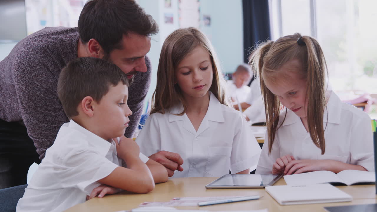 maestro masculino con tres alumnos de escuela primaria vestidos con uniforme usando una tableta digital en el escritorio