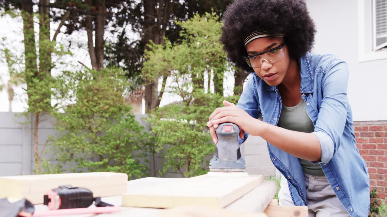 African American woman placing plank then sanding with orbital sander in backyard, smoothing wood