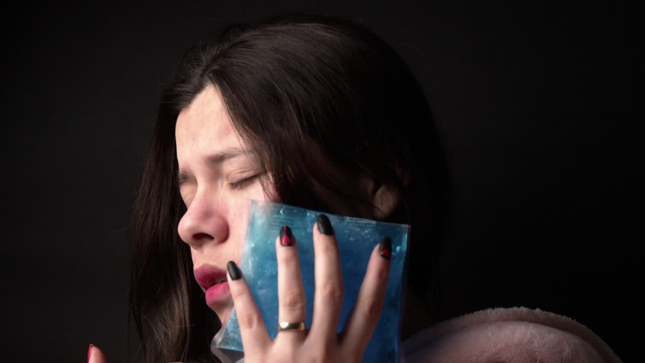 A woman in a soft pink hoodie holds a blue ice pack to her cheek, alleviating toothache pain. Shot captures the emotional relief, with soft lighting enhancing the intimate, close-up scene