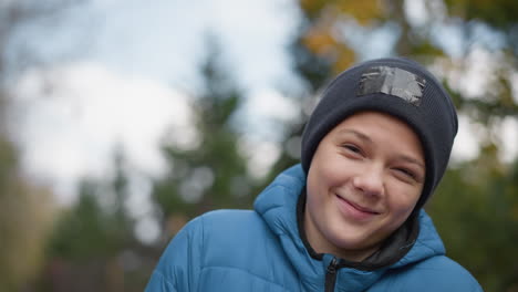 boy wearing black beanie and blue jacket standing outdoors, smiling and enjoying the moment, background features blurred greenery and autumn scenery