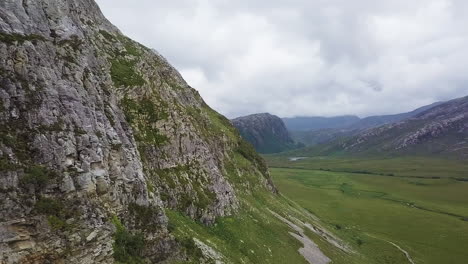 drone flies along rock face and reveals a green valley in the scottish highlands.