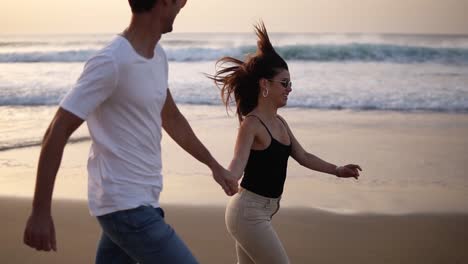 Pareja-De-Vacaciones-Con-Gafas-De-Sol-En-La-Playa-Al-Atardecer-Romántico-Tomados-De-La-Mano-Corriendo-Juguetonamente.-Una-Joven-Pareja-Enamorada-Disfrutando-Del-Romance-Con-Ropa-Informal-Y-Elegante-En-La-Playa.-El-Hombre-Toma-A-Su-Mujer-En-Brazos-Y-Se-Da-La-Vuelta.