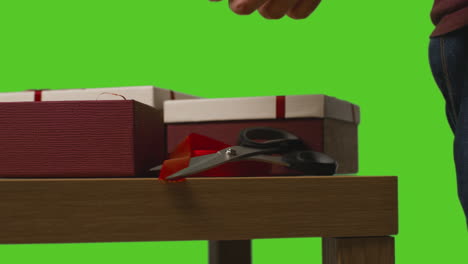 close up of man gift wrapping presents in boxes decorated with ribbon on table shot against green screen