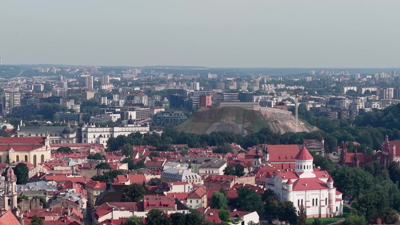 Dynamiske luftopptak glir over Vilnius, og fremhever Gediminas' tårn på toppen av åsen, Gamlebyen med røde tak, historiske kirker og moderne skyline under klart dagslys, ingen mennesker synlige.
