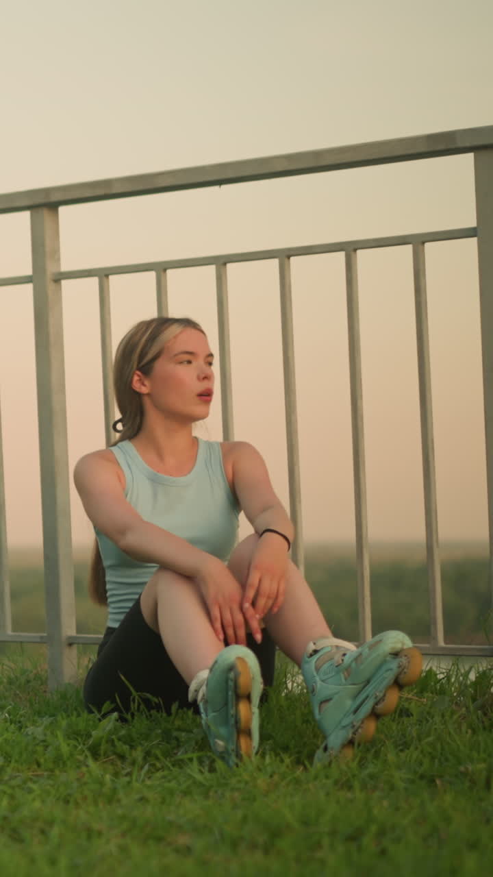 una joven con patines sentada en un campo de hierba cerca de una barandilla de hierro, respirando profundamente con las manos apoyadas en las rodillas, con la suave luz de la tarde y la vegetación en un fondo borroso