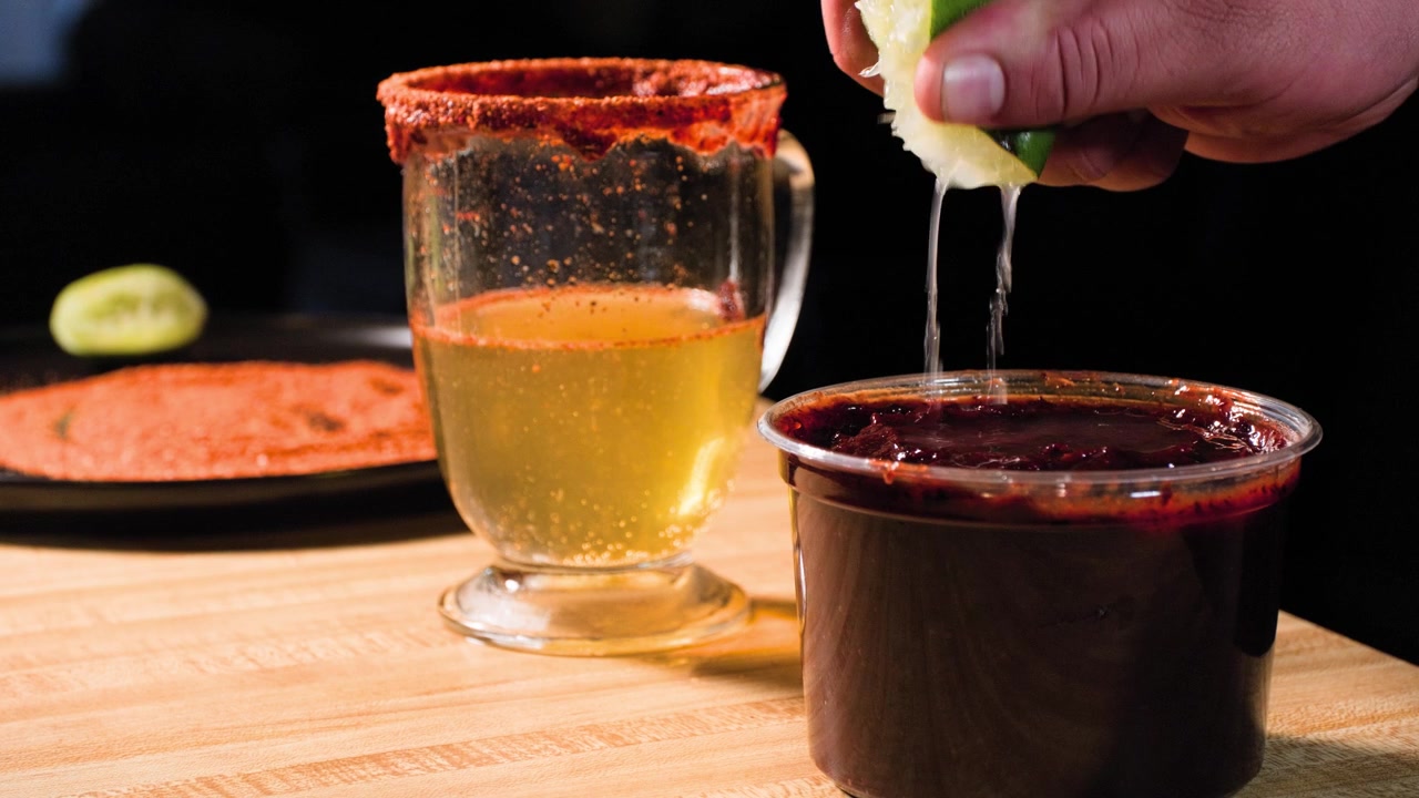 Filmato di cucina in primo piano che mostra un uomo aggiungere lime alla salsa chamoy, con birra e tajin sullo sfondo, mentre prepara da zero una ricetta di salsa chamoy fatta in casa.