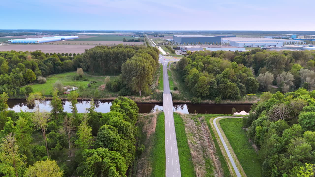 Road leading to the bridge crossing the narrow river. Aerial perspective on the green countryside of the Netherlands. Industrial premises at backdrop.