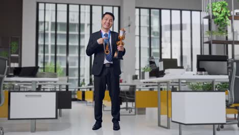 Full Body Of Asian Business Man In A Suit And Tie With A Gold Medal Pointing To A Gold Trophy In His Hand Being Proud Winning As The First Winner In Office