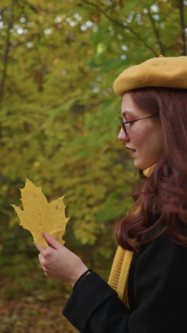 vista lateral de una mujer que lleva una bolsa con una cálida sonrisa sosteniendo una hoja de otoño, observando sus detalles mientras camina por el bosque rodeada de un vibrante follaje dorado, disfrutando de la naturaleza pacífica