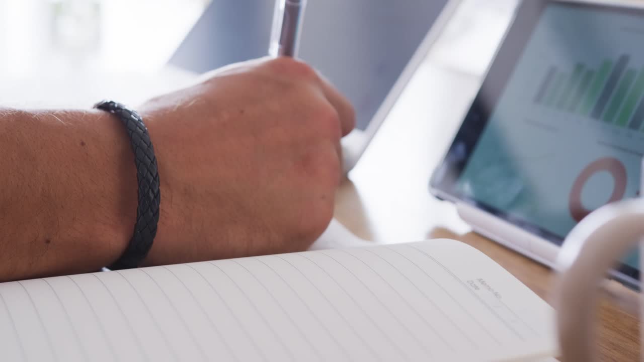 Uomo che consulta grafici su tablet e prende appunti alla scrivania con una penna in un quaderno a righe per studio.