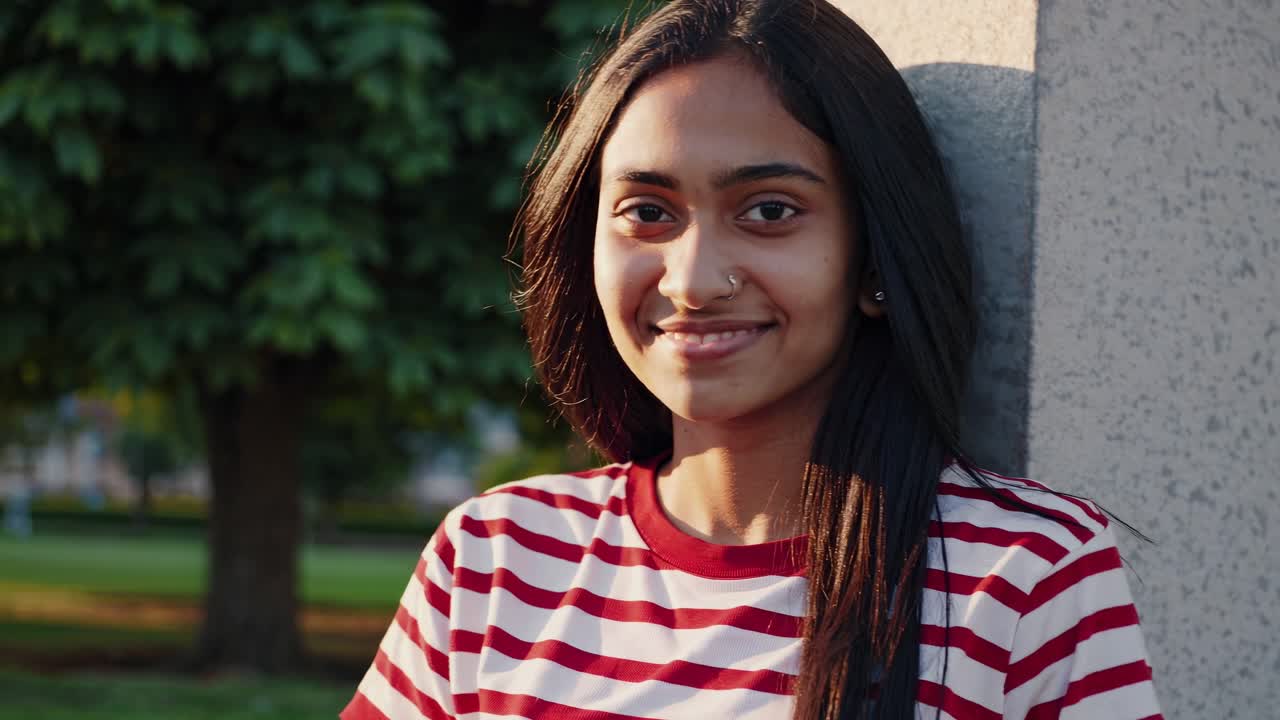 joven segura con cabello largo y oscuro y camiseta a rayas apoyada en una pared en un parque, expresando varias emociones a través de sus expresiones faciales