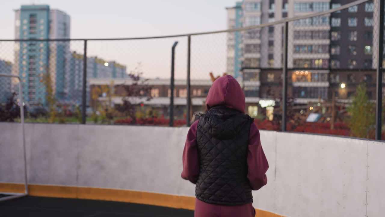 Back view of fitness participant jogging round barbed wire sports court with black quilted vest maroon hoodie curved white barrier on asphalt surface urban tall buildings fence blurred background