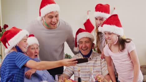 Caucasian-family-wearing-santa-hats-and-face-masks-around-their-necks-using-smartphone-together-in-t