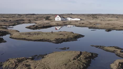 Photographer-stands-on-island-at-small-lake-in-Iceland,-idyllic-cabin-in-background,-aerial