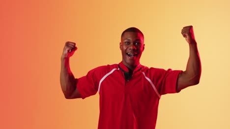 Portrait-of-happy-african-american-male-rugby-player-raising-hands-over-orange-lighting