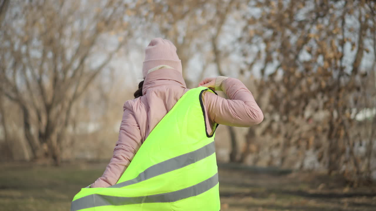 Woman in pink winter jacket and beanie viewed from behind putting on bright yellow reflective safety vest outdoors in park with bare trees, preparing for activity or volunteer work in cold weather