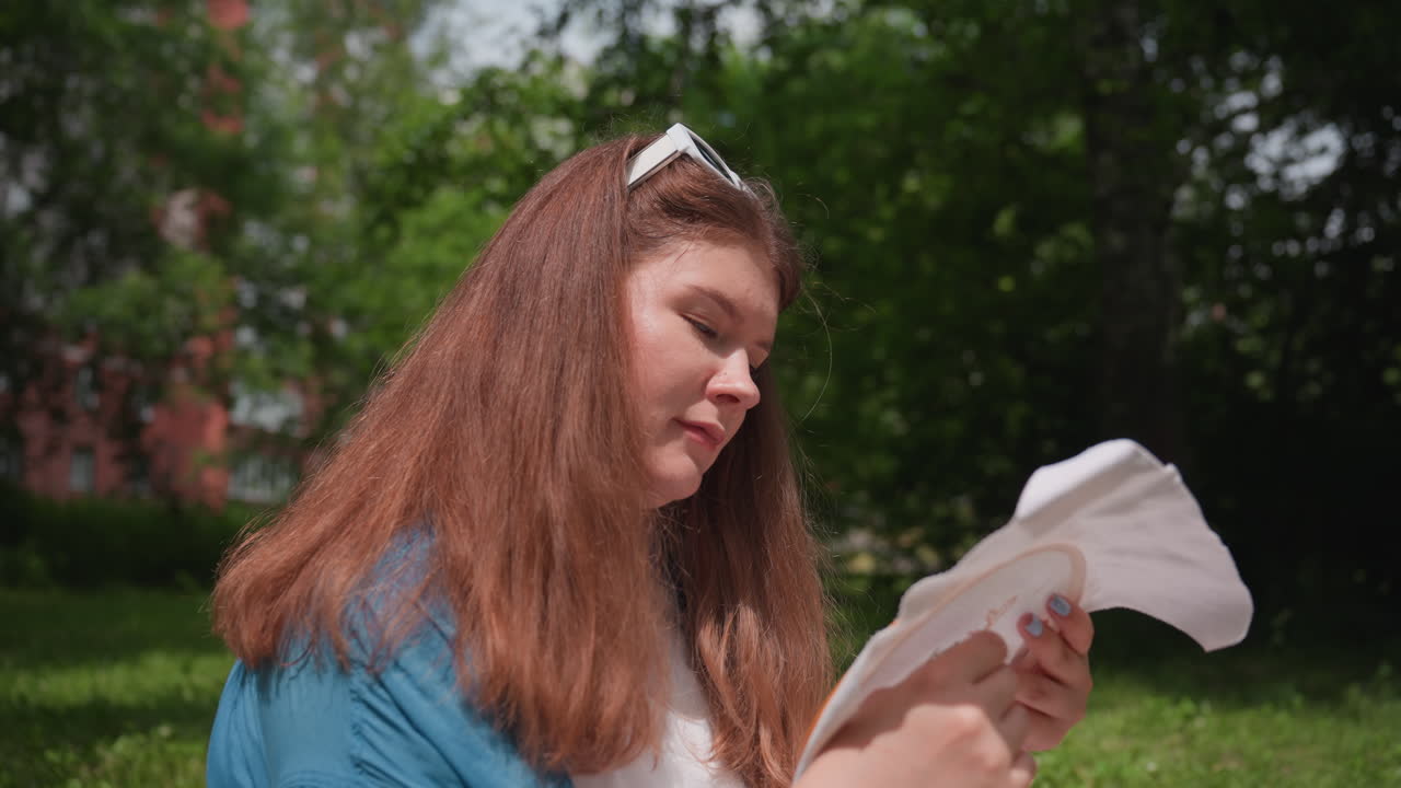 Elegant lady seated on grass near residential building, embroidering fabric hoop with focus and calmness during bright sunny day, expressing creative passion