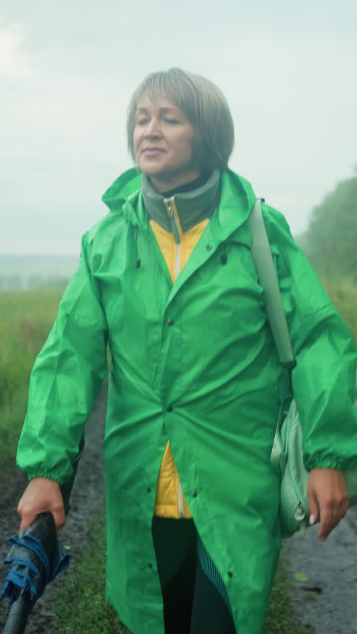 Woman in green raincoat carrying mint-colored bag and umbrella, walking along a muddy path surrounded by greenery and trees under a misty sky, looking into the distance