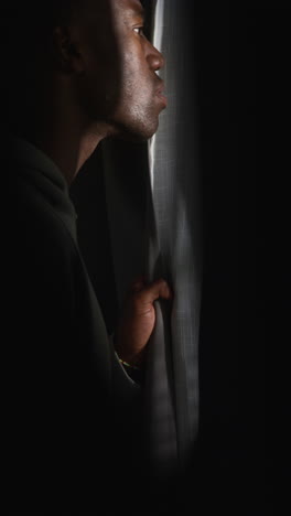 vertical video of unhappy and depressed young man at home looking out of window suffering with agoraphobia or paranoia 3