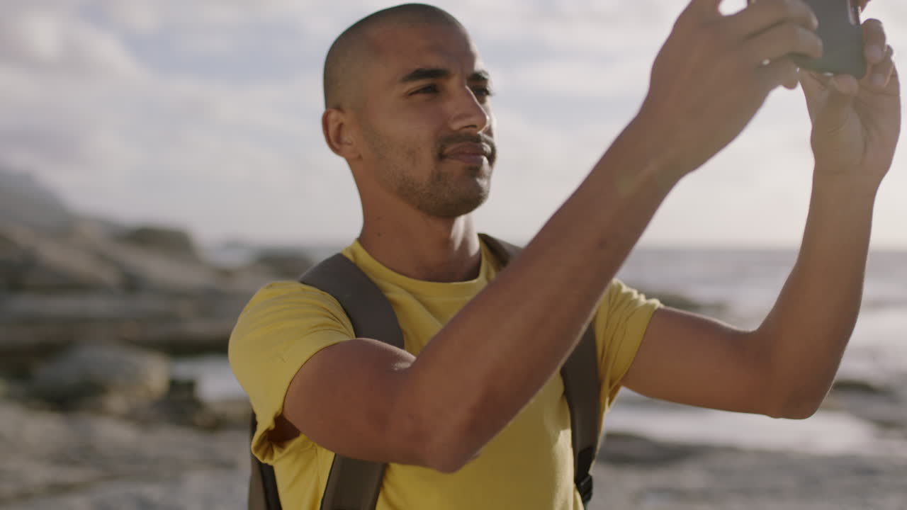 joven hombre hispano atractivo tomando una foto en la playa usando el teléfono sonriendo feliz