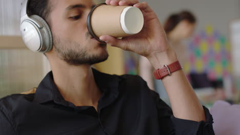 Cerrar-A-Un-Joven-Hispano-Usando-Una-Computadora-Portátil-Navegando-Mensajes-En-Línea-Compartiendo-Comunicación-En-Red-Estudiante-Tomando-Café-Disfrutando-Escuchando-Música-En-Una-Oficina-Moderna