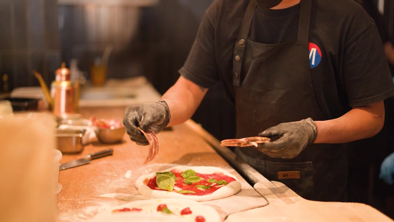 Chef con guanti neri posiziona il prosciutto sull'impasto della pizza con salsa di pomodoro e basilico.