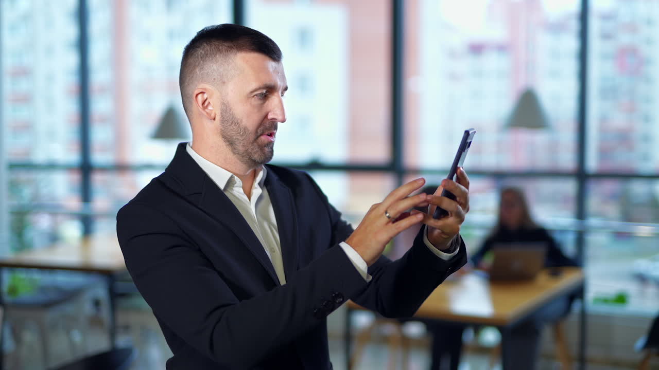 Happy confident smiling man in suit takes selfie in office. Female employees sitting at desk at backdrop in blur.