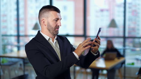 happy confident smiling man in suit takes selfie in office. female employees sitting at desk at backdrop in blur.