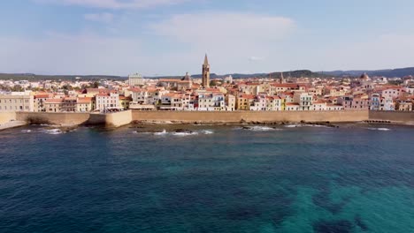 Clip-De-Drones-Sobre-El-Océano-Azul-Con-La-Pintoresca-Ciudad-De-Alghero-En-Cerdeña,-Italia-Que-Se-Extiende-A-Lo-Largo-Del-Fondo