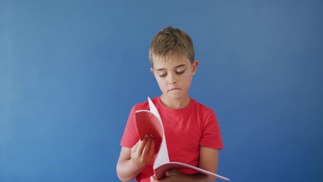 Retrato-De-Un-Niño-Caucásico-Feliz-Sosteniendo-Un-Libro-En-La-Cabeza-Sobre-Fondo-Azul,-Cámara-Lenta