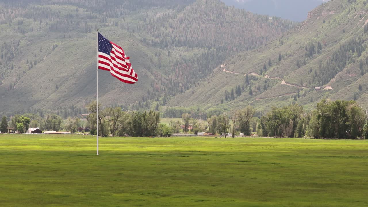 Bendera Amerika besar berkibar tertiup angin di atas padang rumput hijau, rumah peternakan, dan jalur pegunungan, dengan Pegunungan San Juan di latar belakang