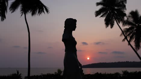 Statuensilhouette-Vor-Einem-Lebhaften-Tropischen-Sonnenuntergang-Mit-Palmen-Am-Meer