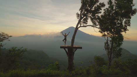 woman balancing on one leg doing yoga tree pose on wood platform, mount agung, sunset