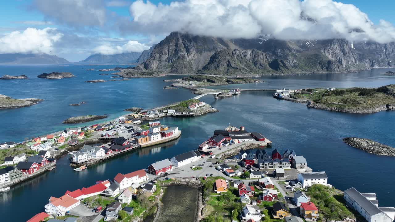 Aerial flight over fishing village of Henningsvaer, road leading to Festvagtinden Mountain in distance, Lofoten, Norway