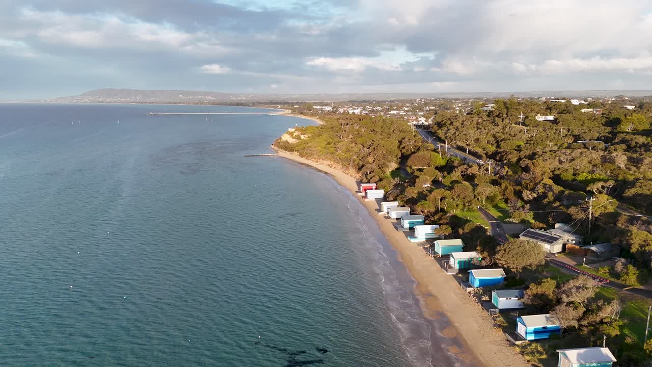 Drone glides above vibrant bathing boxes, sandy beach, and coastal vegetation in morning sunlight