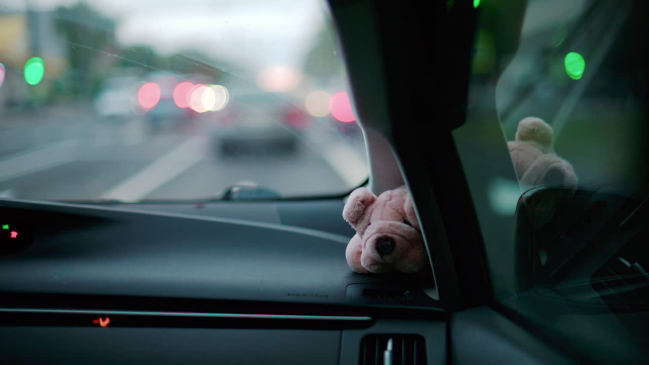 A small stuffed toy rests on a car dashboard as blurred city lights pass by through the windshield
